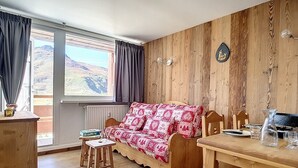 Una habitación acogedora con pared de madera, un sofá de madera con una manta de motivos rojos y blancos, una mesa de comedor de madera con sillas y una ventana con cortinas.