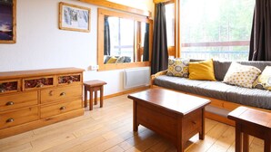 Ein gemütliches Zimmer mit einem Holzschrank, einer Couch mit gemusterten Kissen, einem Holztisch und großen Fenstern mit Vorhängen.