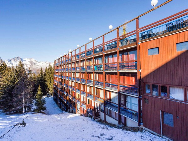 Un hotel de varios pisos con balcones, una pista de esquí al fondo y nieve en el suelo.