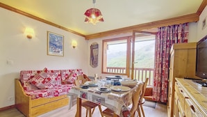 A cozy dining area with a wooden table set for four, a red and white patterned sofa, and a television on a wooden cabinet. - 2-room apartment · Ski-in/Ski-out · Southwest balcony (Les Menuires)