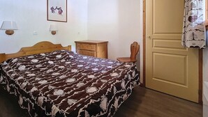 A bedroom with a bed, a wooden dresser, a chair, a door, and a wall-mounted lamp. - 2-room apartment · Ski-in/Ski-out · Southwest balcony (Les Menuires)
