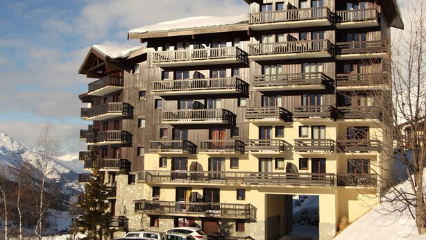 A multi-story hotel with balconies, a stone base, and a snowy mountain backdrop.