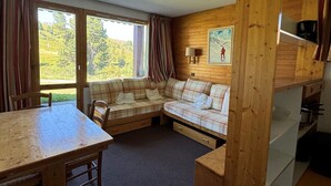 Ein gemütlicher Raum mit einer hölzernen Bank, einem karierten Sofa, einem Esstisch und einem großen Fenster mit Blick auf Grünflächen.