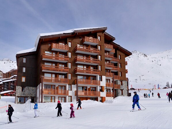 A multi-story building with balconies, likely a hotel or resort, surrounded by snow and skiers.