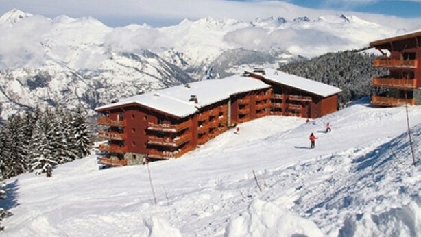 Een houten chalet met meerdere balkons, omgeven door besneeuwde bergen en bomen.