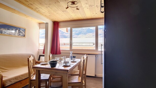 Um quarto acolhedor com teto de madeira, uma mesa de jantar posta com pratos, xícaras e um vaso, e uma cama com cortina vermelha.