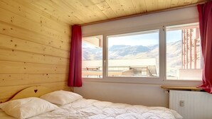 Quarto com cama, paredes de madeira, janela com vista para montanhas e radiador.