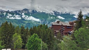 Paisaje montañoso con vistas a picos nevados y un valle en la distancia. En primer plano, edificios con balcones, rodeados de árboles.