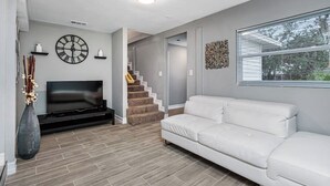 Uma sala de estar moderna com um sofá branco, uma TV de tela plana, um relógio decorativo e uma escada que leva a outro andar.