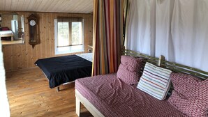 A wooden cabin with a bed, a small table, a clock, and a window with curtains. - Maison au Calme sur Jardin Arboré (Saint-Georges-d'Oléron)