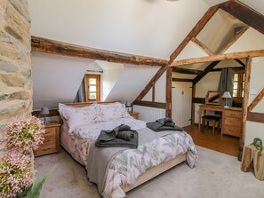 Et soveværelse med skråvæg, seng med blomstret sengetøj, skrivebord med stol og et vindue med gardiner.