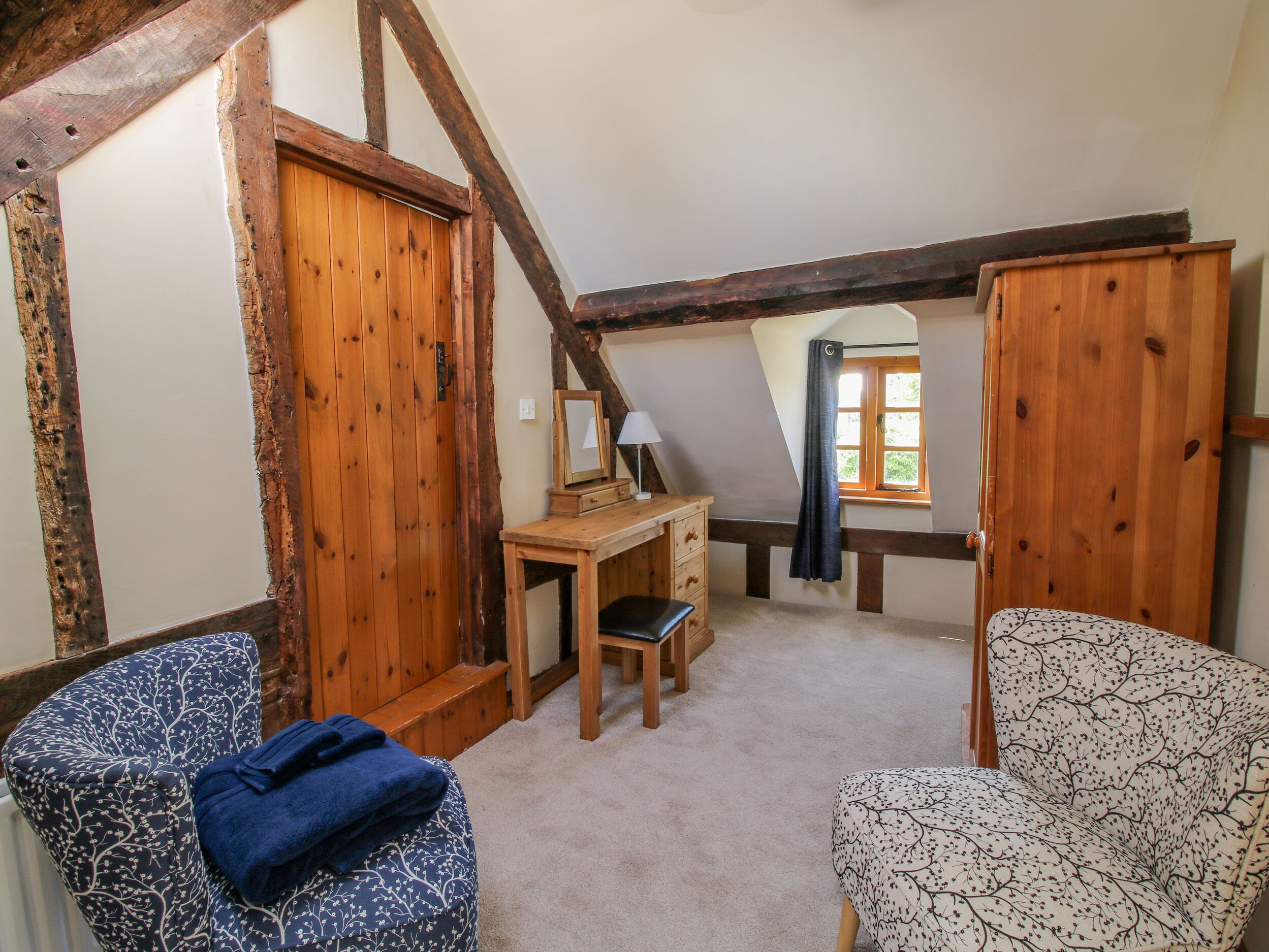 Ein gemütliches Schlafzimmer im Dachgeschoss mit freiliegenden Holzbalken, einem Holztisch mit Spiegel, zwei gemusterten Stühlen und Blick durch ein Fenster mit Vorhang.
