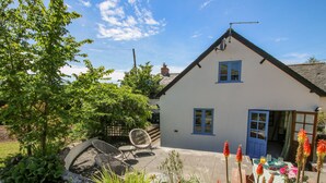 A white house with blue trim, a garden with various plants, and a patio area with outdoor furniture. - 46-47 Lydbury North (Lydbury North)