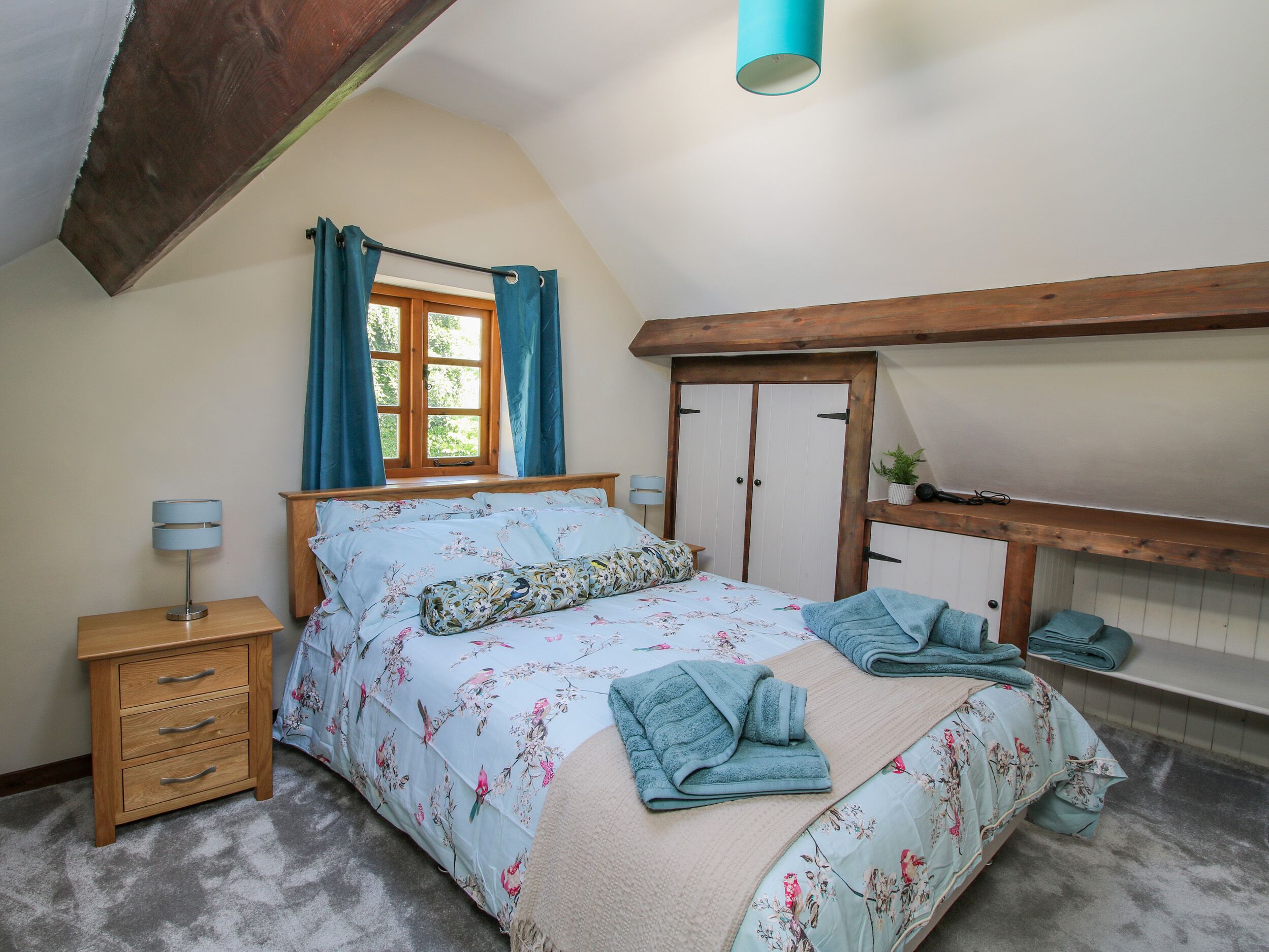 A bedroom with a bed, a nightstand, a window with curtains, a wooden ceiling, and a wardrobe.