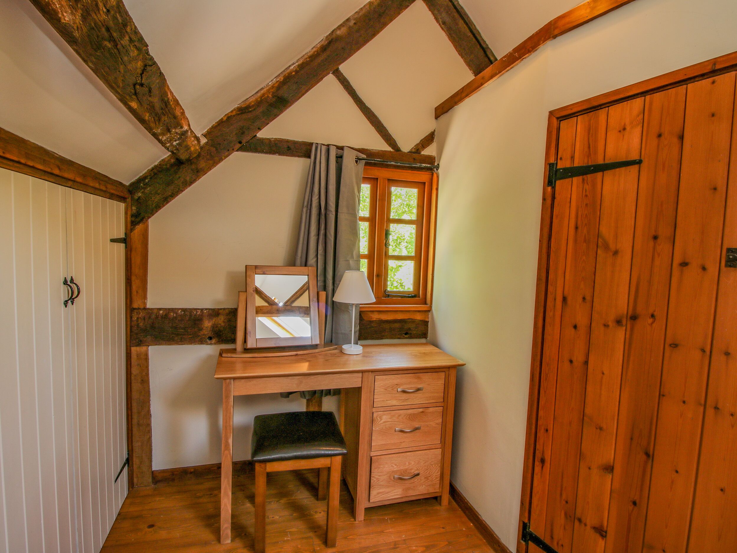 Una stanza con soffitto spiovente, travi in legno, una scrivania con cassetti, una sedia, una finestra con tende e una porta in legno.