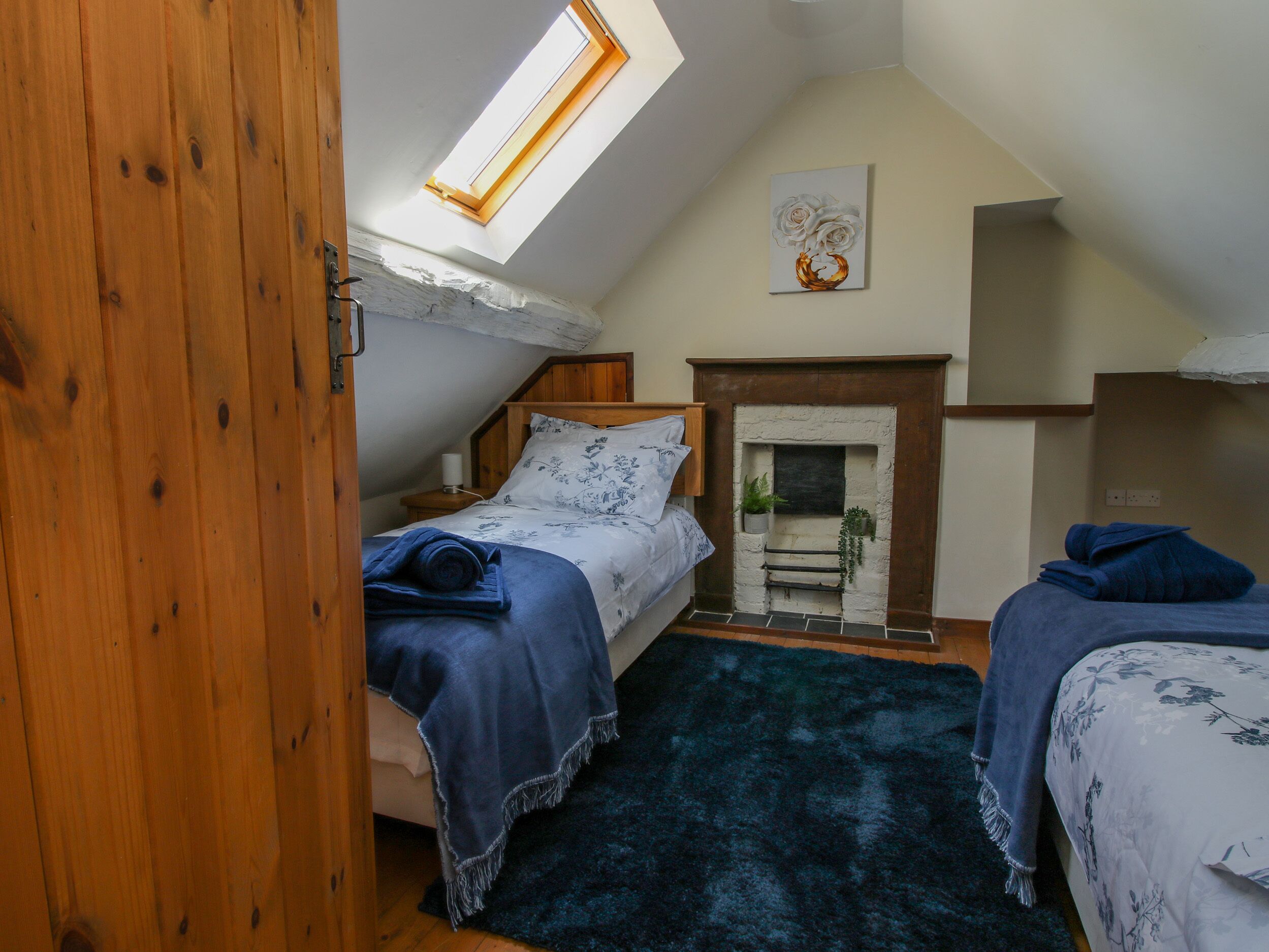 Une chambre confortable avec un plafond en pente, une lucarne, une cheminée et un lit recouvert d’une couverture bleue.