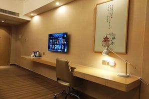 A hotel room with a wooden desk, a chair, a flat-screen TV mounted on the wall, a lamp, and a decorative wall piece. - Xin Zhenhao Business Hotel (Shantou)