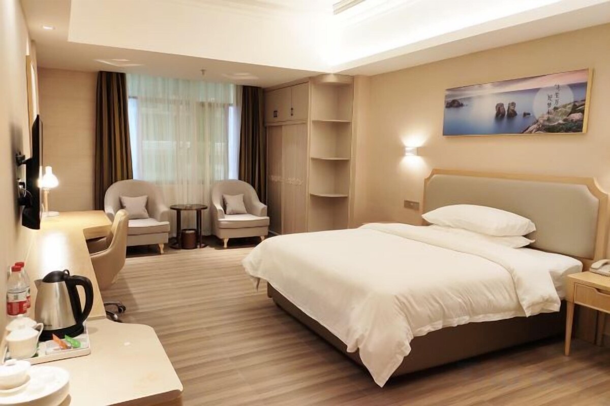 Una habitación de hotel con una cama grande, un escritorio con lámpara, dos sillones, una mesita y un cuadro en la pared.