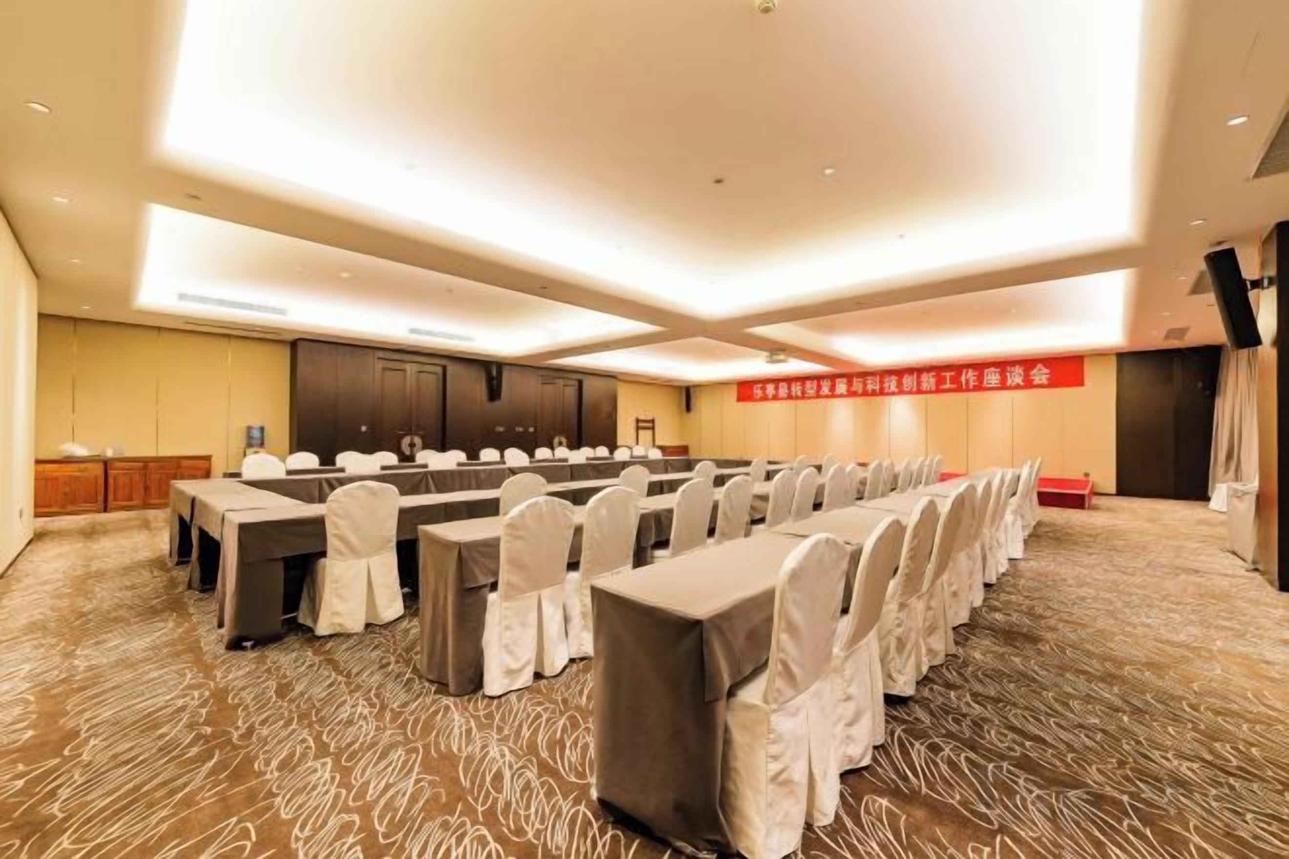 Un salón de conferencias con filas de mesas y sillas, un atril y piso alfombrado.