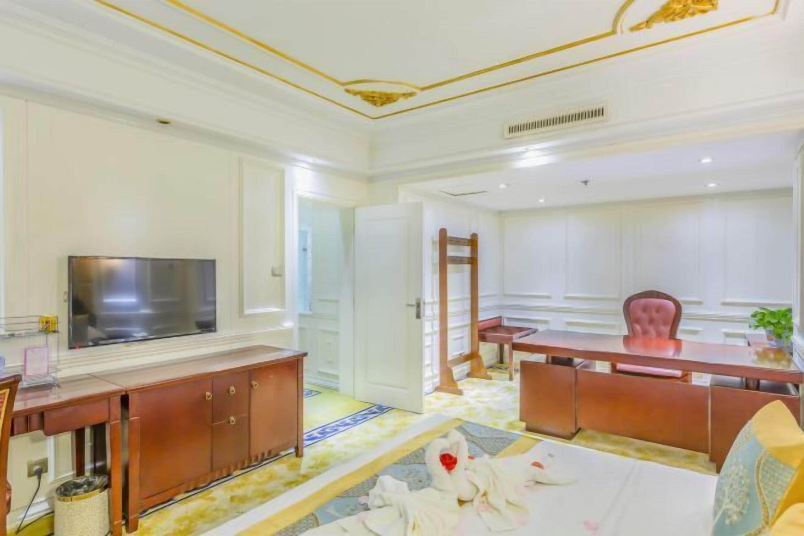 A hotel room with a large flat-screen TV, a wooden cabinet, a dining table with chairs, and a carpeted floor.