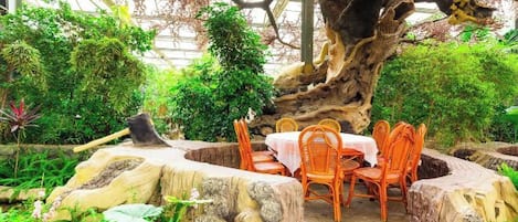 Un jardín interior con zona de comedor, con una mesa y sillas sobre una base de piedra, rodeado de abundante vegetación y un gran tronco de árbol.