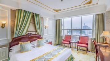 A hotel room with a large bed, two chairs, a desk, and a view of the city through the window.