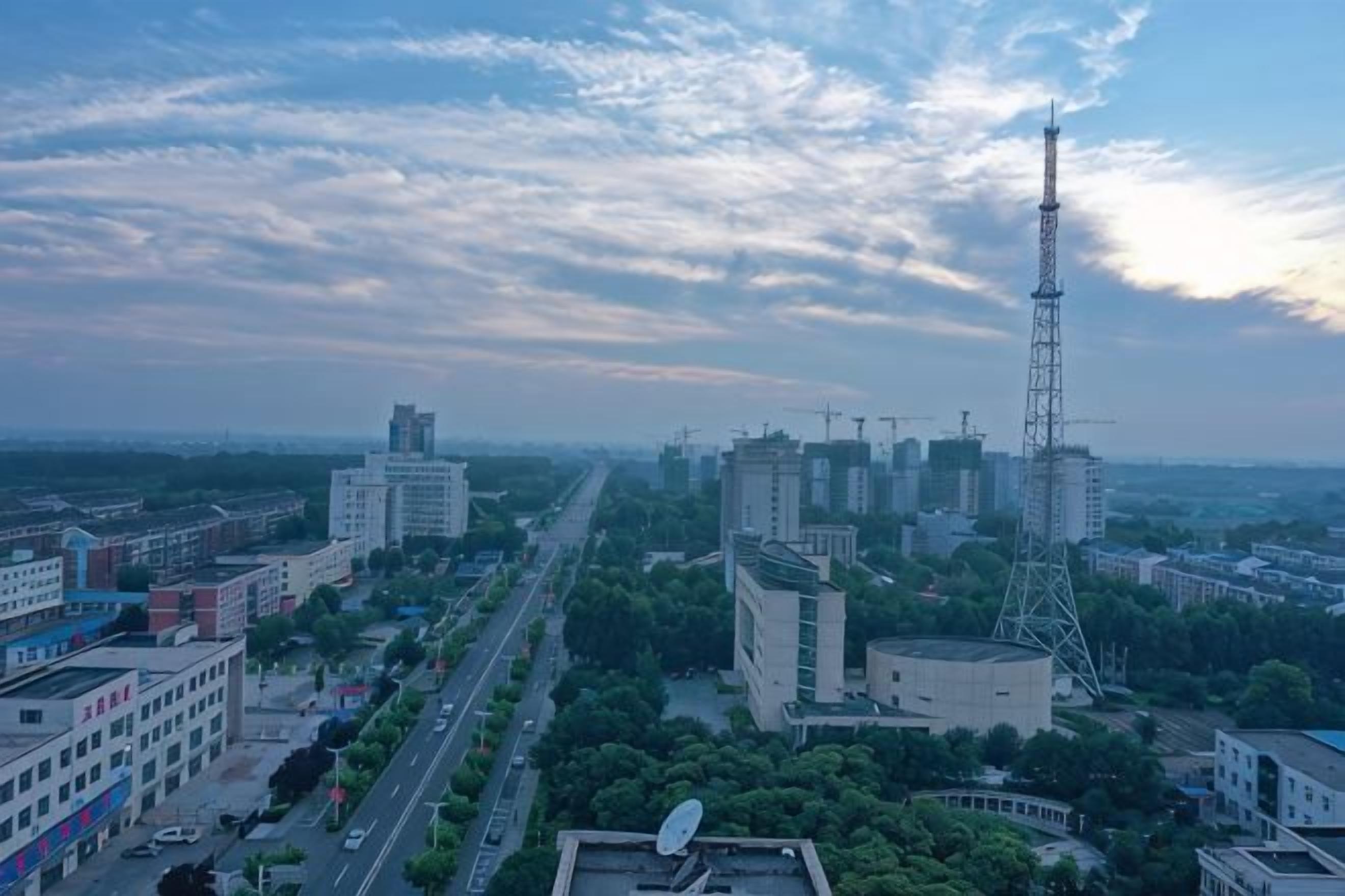 A cityscape with modern buildings, a prominent communication tower, and a road lined with trees.