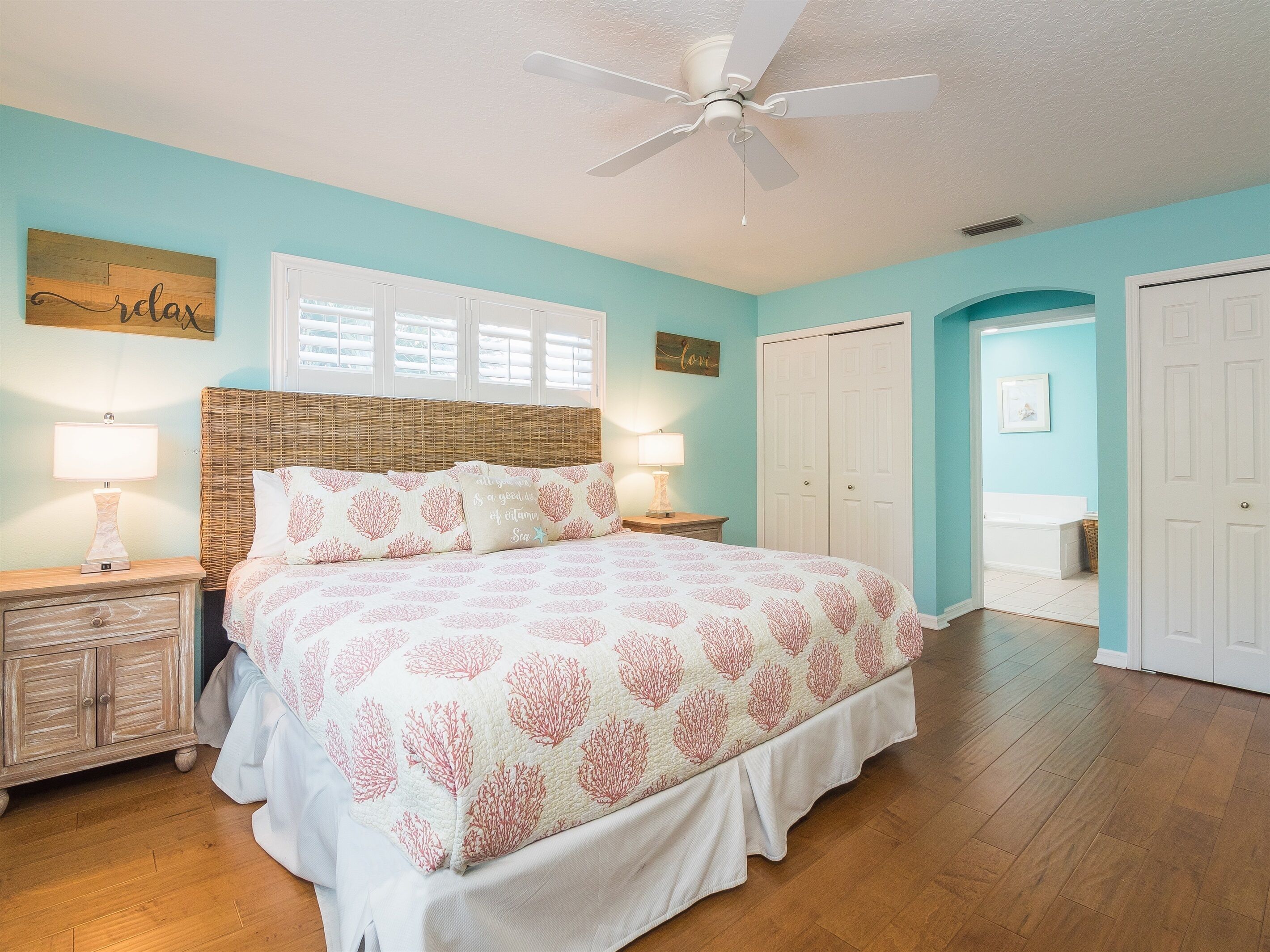 A bedroom with a bed, two bedside tables, a ceiling fan, and a door leading to another room.