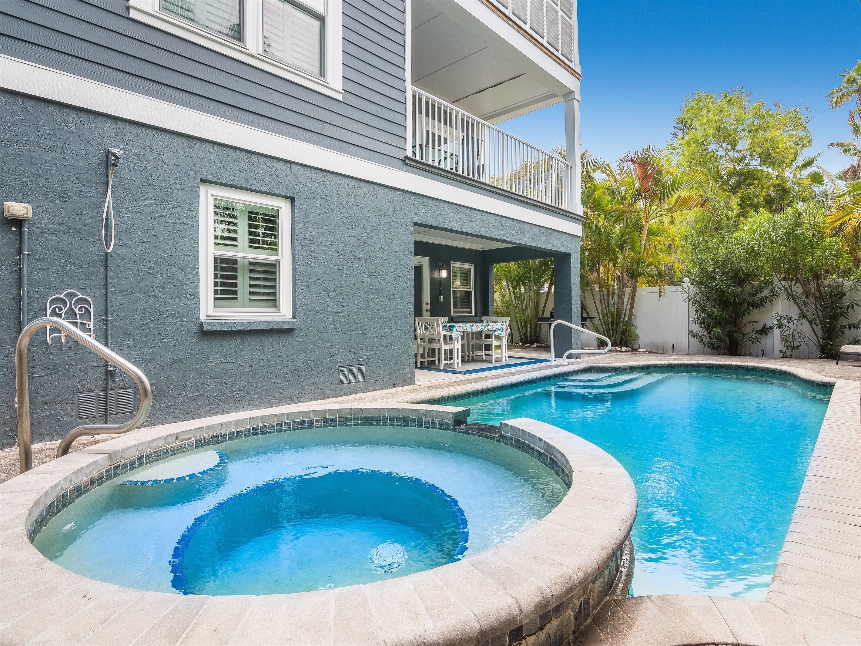 A swimming pool with a hot tub, surrounded by a house with a balcony and outdoor furniture.