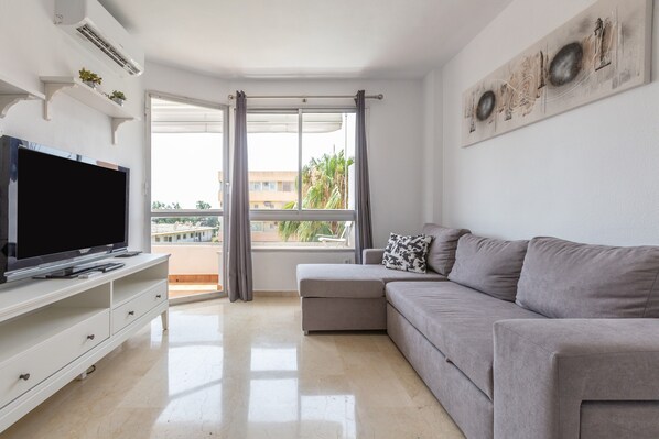 A modern living room with a flat-screen TV, a grey sofa, a white entertainment unit, and a large window with curtains.
