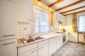 Une cuisine avec des meubles blancs, un évier à deux bacs, une cuisinière et une fenêtre avec un rideau.