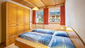 A wooden bed with blue bedding, a wooden wardrobe, a window with red curtains, and a view of houses. - Pet-Friendly Holiday Apartment 'Kuenz Dolomiten Drau' with Mountain View (San Candido)