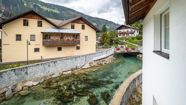 A building with a balcony overlooking a clear water body, a stone wall, and a mountainous backdrop. - Pet-Friendly Holiday Apartment 'Kuenz Dolomiten Drau' with Mountain View (San Candido)