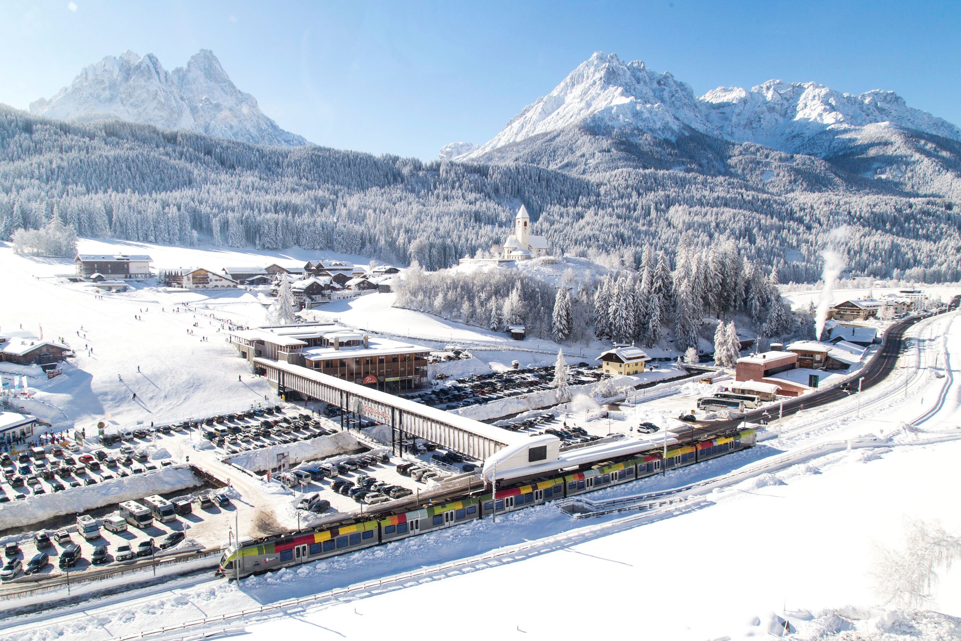 Una estación de tren nevada con un tren, edificios y montañas al fondo.