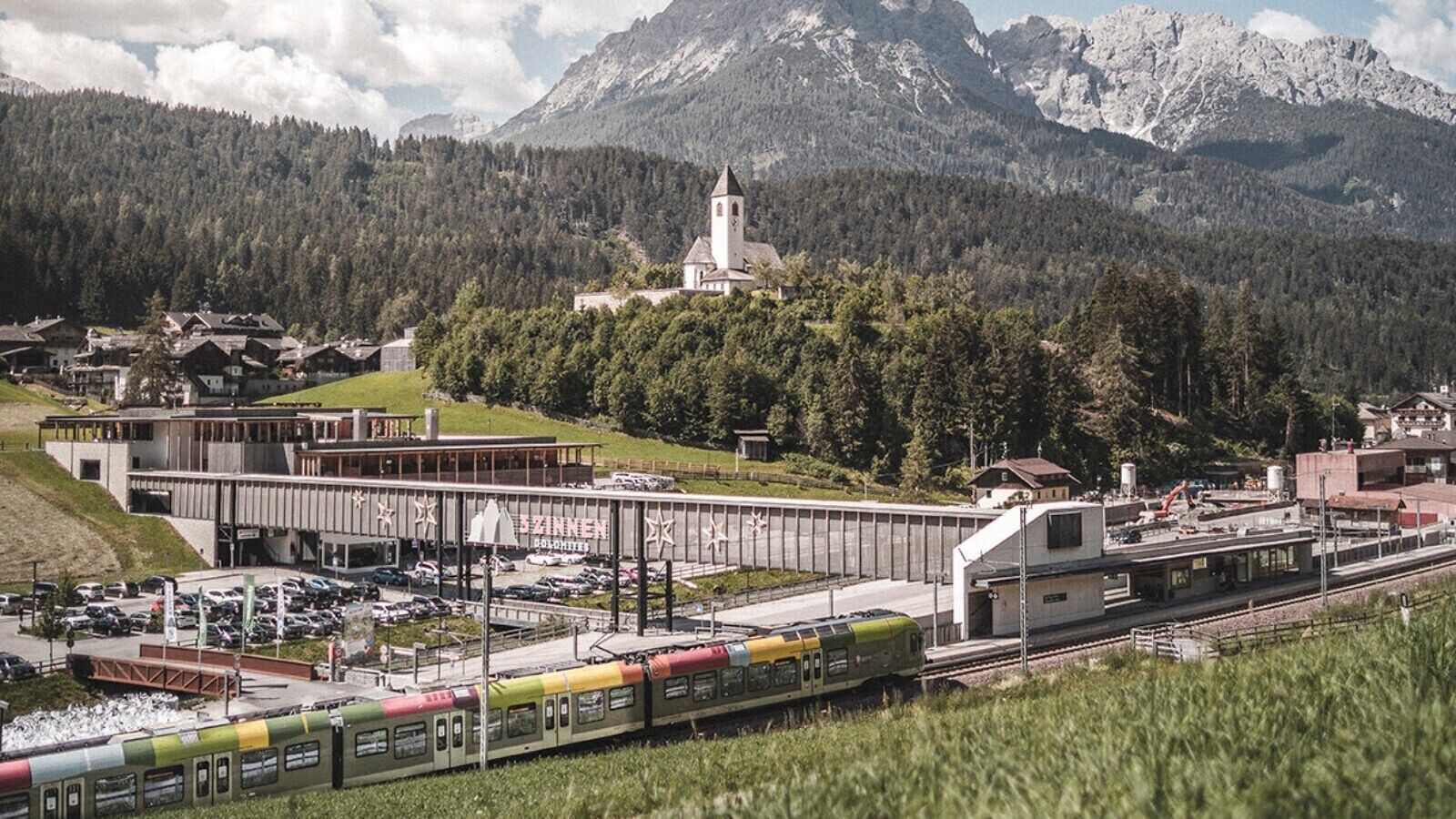 Ein Bahnhof mit einer Brücke, einer Kirche und Bergen im Hintergrund.