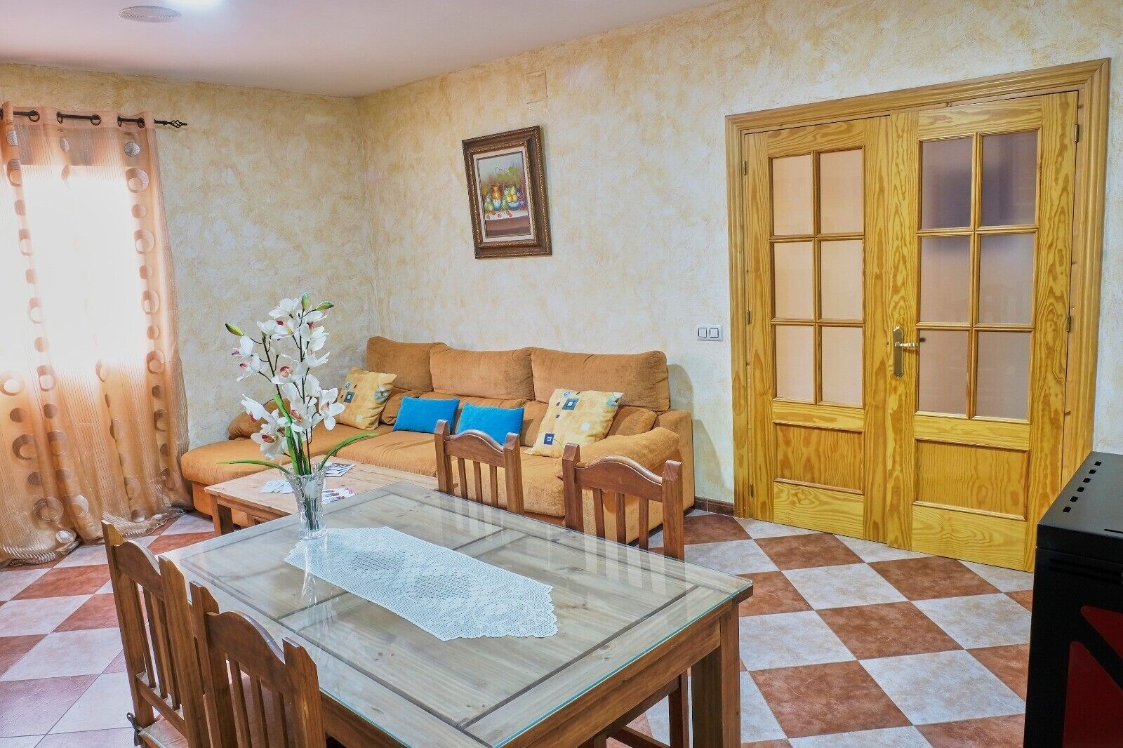 Una habitación con puerta de madera, un sofá con cojines, una mesa de vidrio con sillas y un cuadro en la pared.