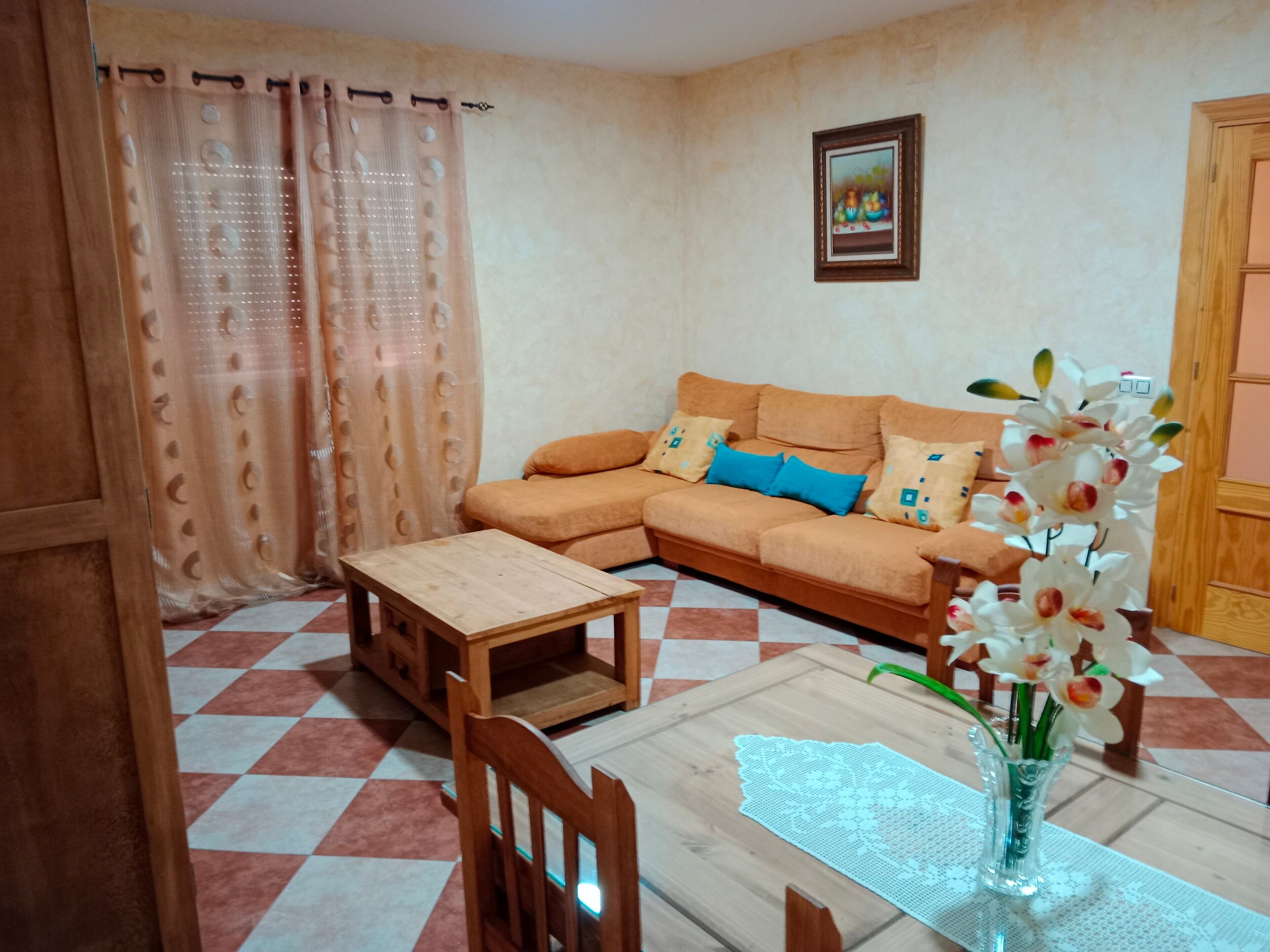 Una sala de estar con un sofá, una mesa de madera, una silla, un florero con flores y un piso a cuadros.