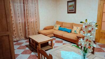 Una sala de estar con un sofá, una mesa de madera, una silla, un florero con flores y un piso a cuadros.