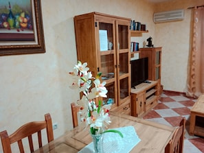 Un coin repas comprenant une table, des chaises, un vase avec des fleurs, un buffet en bois avec des portes en verre et un téléviseur.