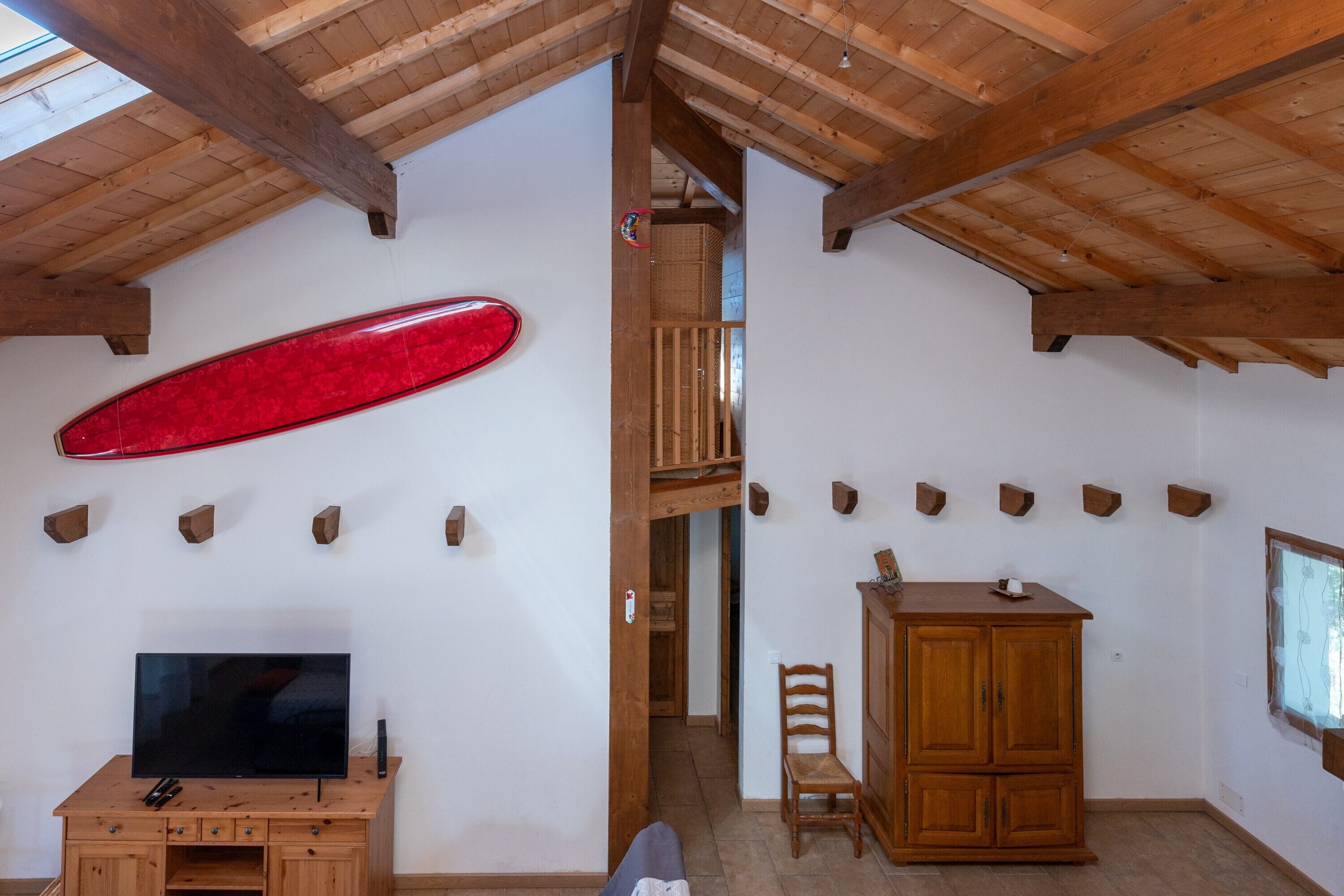 Ein Zimmer mit Fernseher auf einem Holzschrank, einem roten Surfbrett an der Wand und einer Holzbalkendecke.