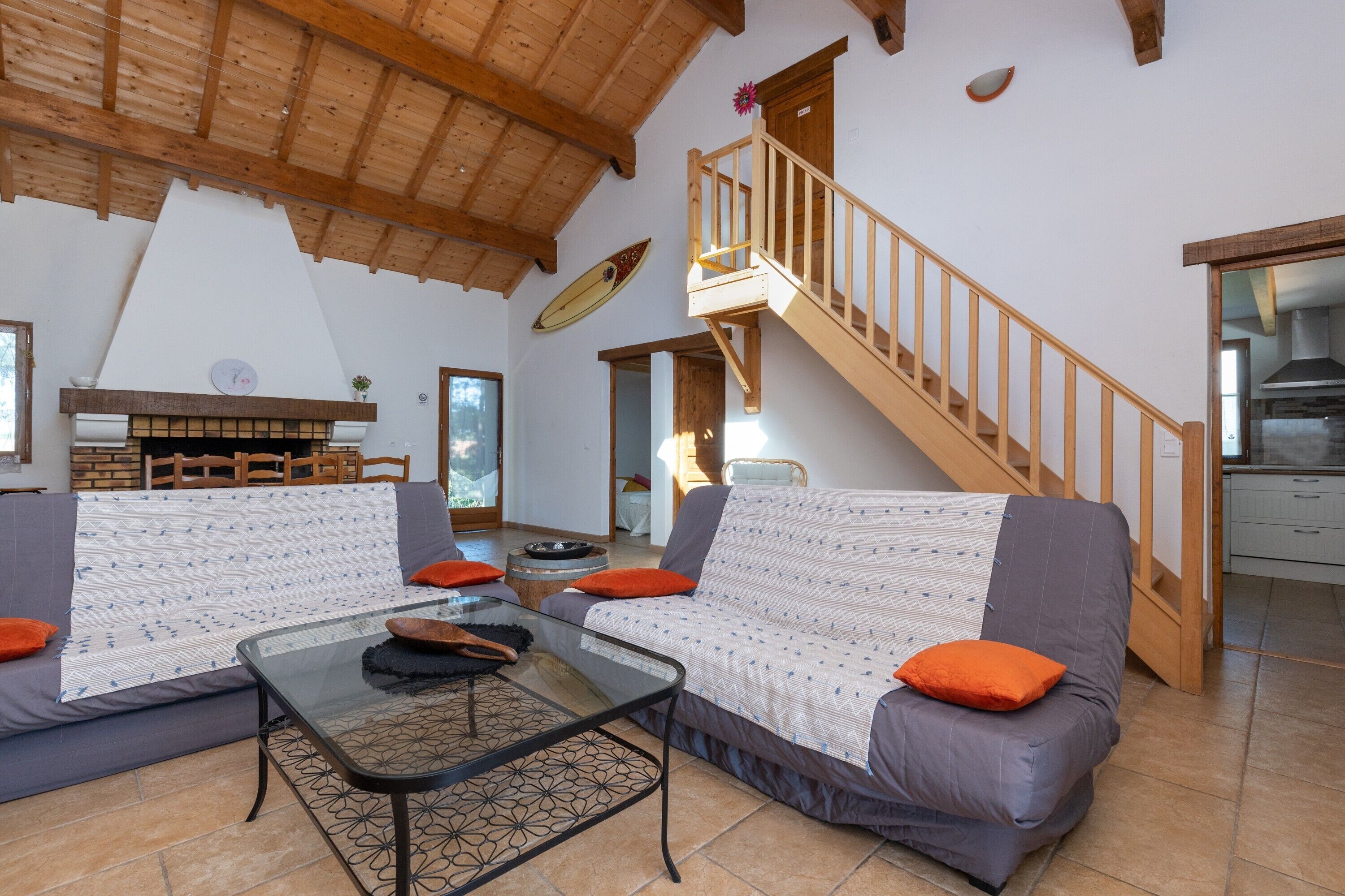 A living room with a sofa, a glass coffee table, a fireplace, a wooden staircase, and a kitchen area.