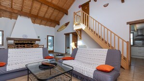 Una sala de estar con sofá, una mesa de centro de vidrio, chimenea, una escalera de madera y una zona de cocina.
