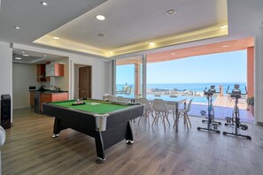 A modern living room with a pool table, exercise equipment, and a view of the sea.