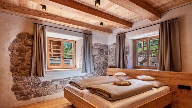 Un letto di legno in una stanza con pareti in pietra e travi in legno, ampie finestre con tende e un camino in pietra.