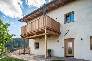 Un edificio de dos pisos con un balcón de madera, piso de piedra y vista a un área boscosa.