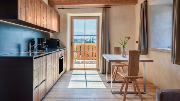 Una cocina moderna con armarios de madera, horno empotrado y cafetera. Cuenta con un comedor con mesa y sillas, y vistas a una terraza de madera a través de una puerta de vidrio.