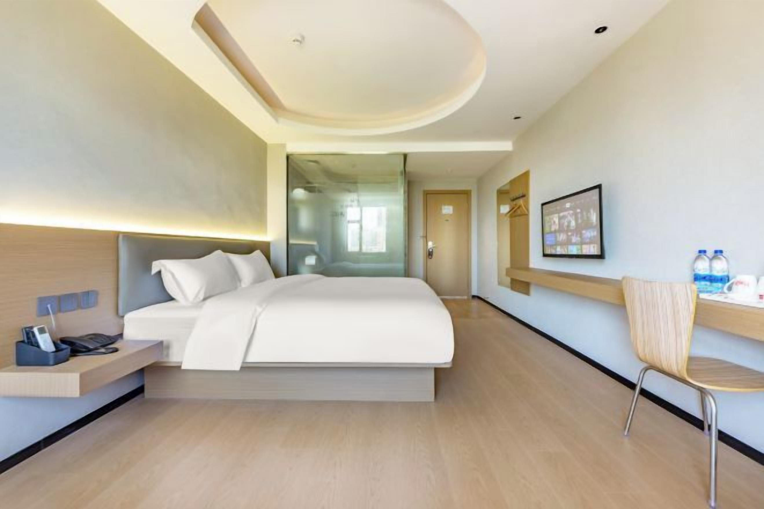 A modern hotel room with a bed, bedside table, chair, and a view of the hallway.