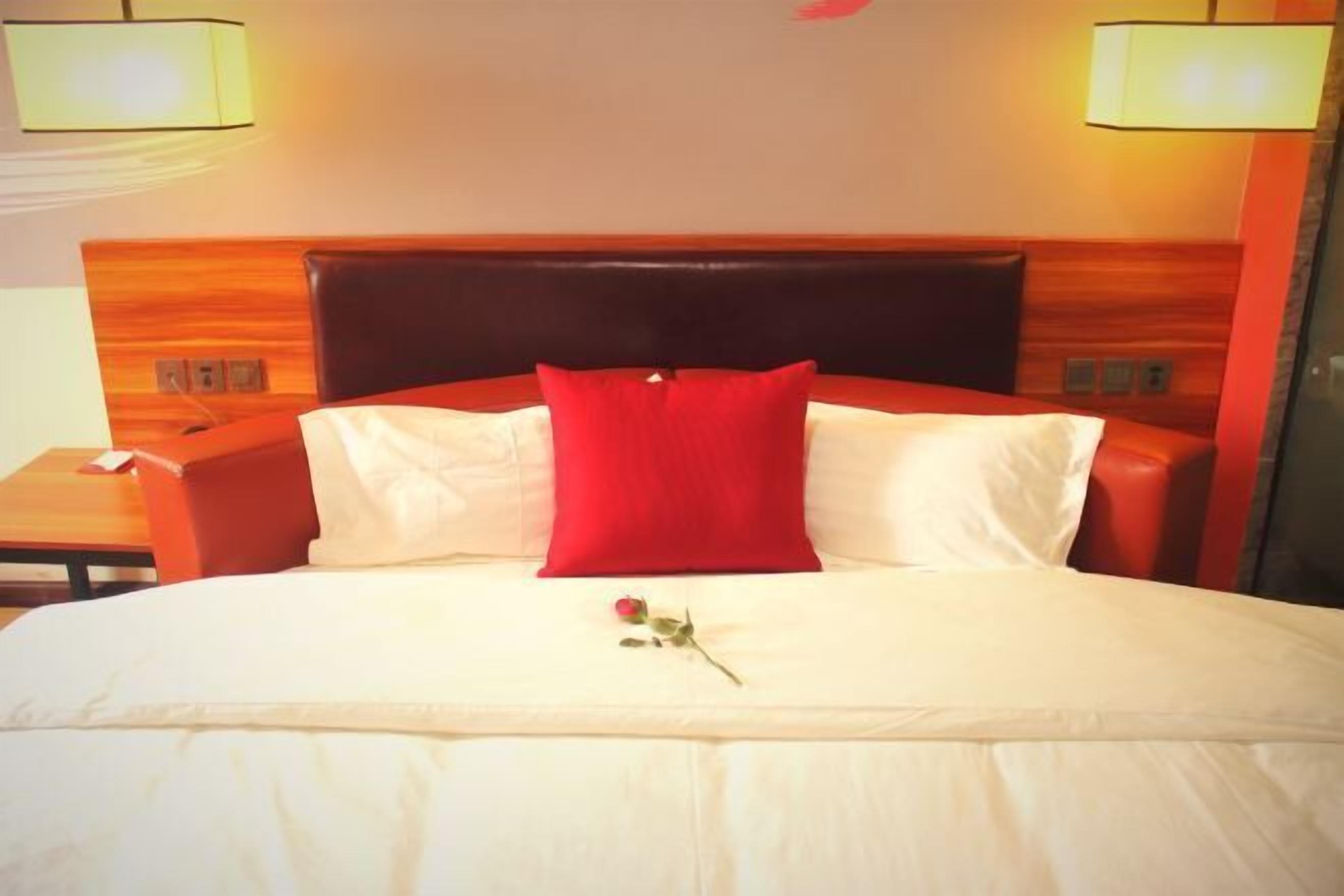 A hotel room with a large bed, a wooden headboard, two bedside lamps, and a red pillow.