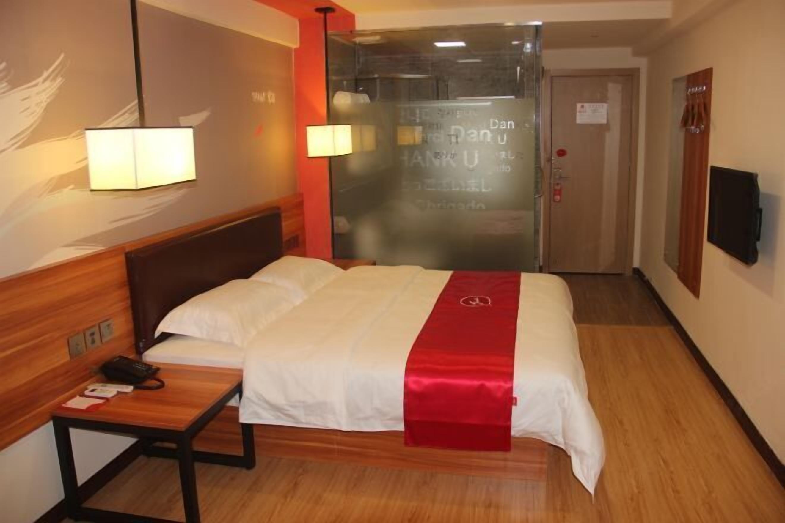 A hotel room with a bed, bedside table, lamp, and a glass door with a sign.
