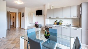 Une cuisine moderne avec des meubles blancs, un téléviseur à écran plat et un coin repas comprenant une table en verre et des chaises bleues.